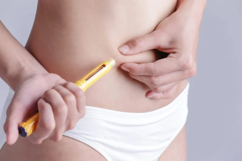 A person administering an injection into their abdomen with a yellow pen-like device.